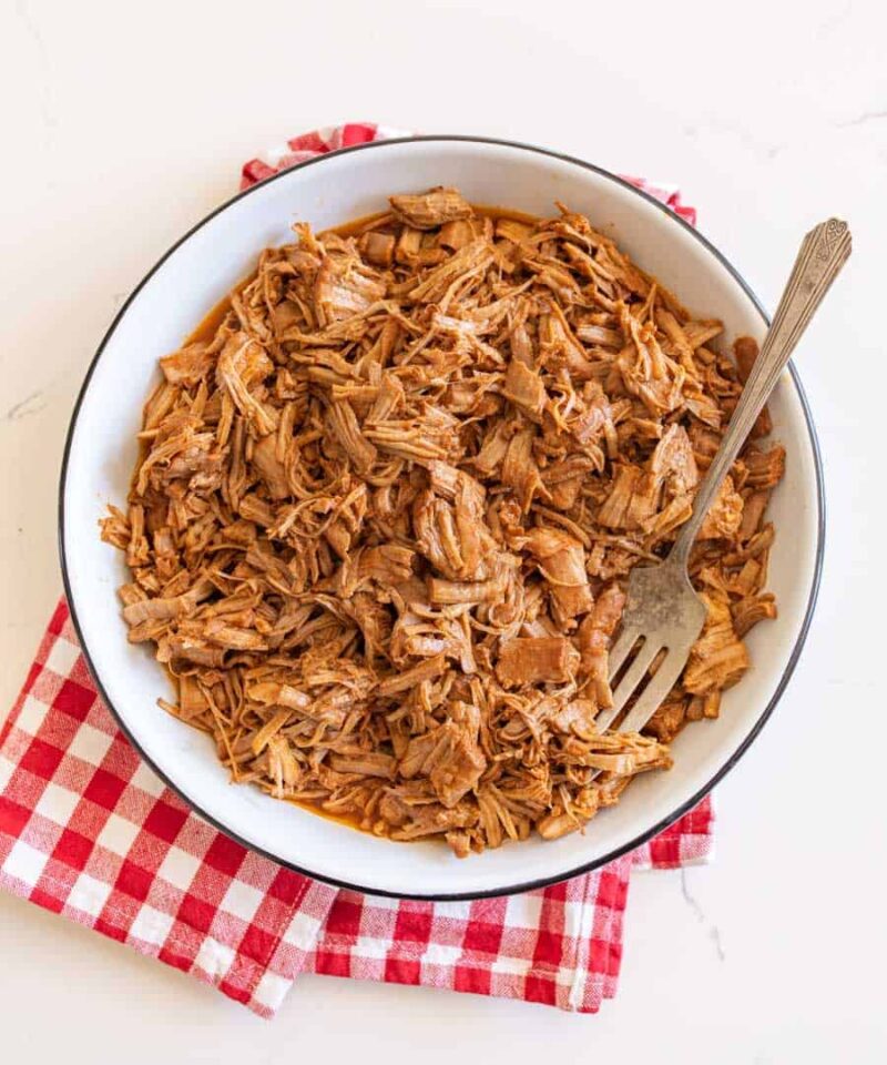 A white bowl filled with shredded Instant Pot pulled pork tenderloin sits on a red and white checkered cloth, with a fork resting inside the bowl.