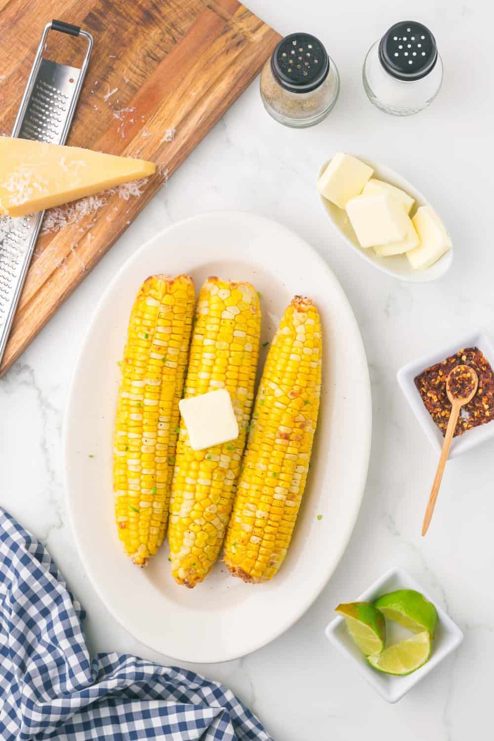 Three grilled corn on the cob with a pat of butter on top are served on a white plate. Surrounding items include butter, cheese, chili flakes, lime wedges, salt, pepper, and a blue checkered napkin—perfect for air fryer corn on the cob in husk.