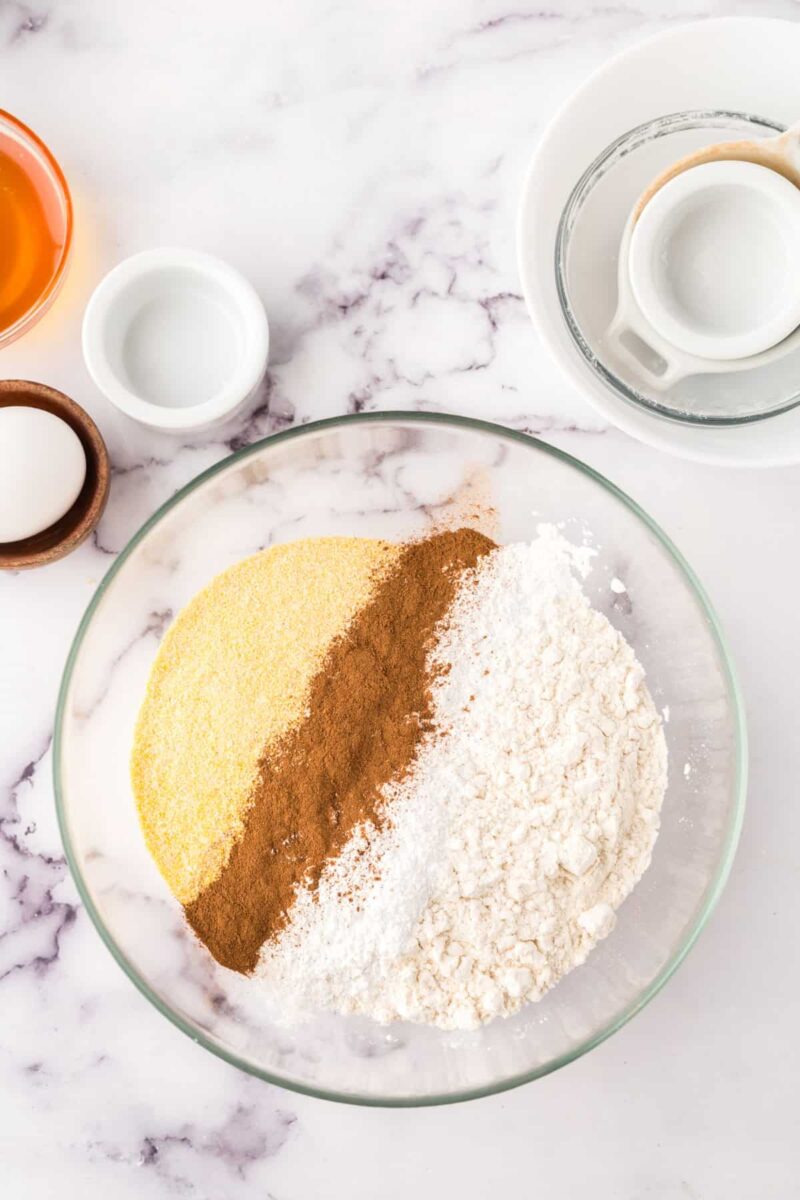 A glass bowl containing lines of cornmeal, cinnamon, and flour sits on a marble countertop—ingredients ready to be mixed for delicious pumpkin corn muffins, surrounded by small bowls with various baking essentials.
