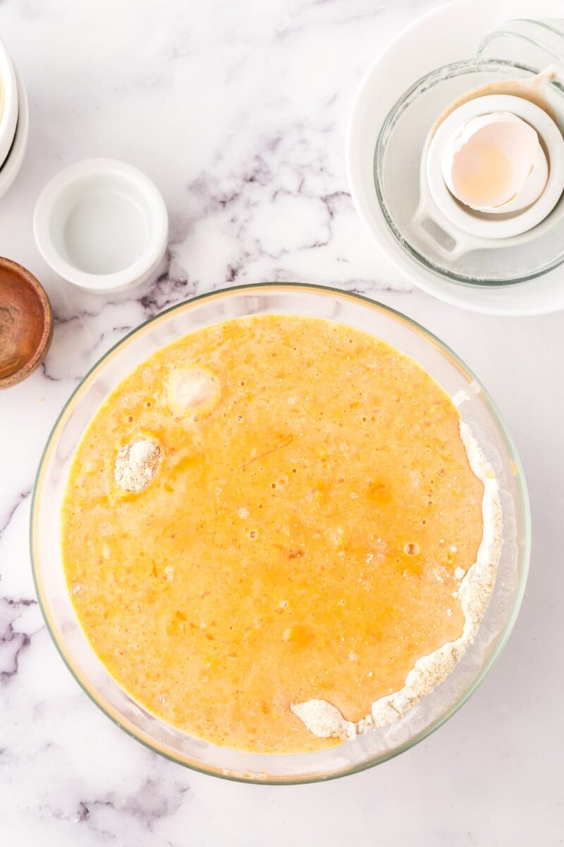 A large glass bowl filled with yellow, partially mixed pumpkin corn muffin batter sits on a marble countertop. Nearby are a cracked egg in an egg separator, small bowls, and a wooden dish.