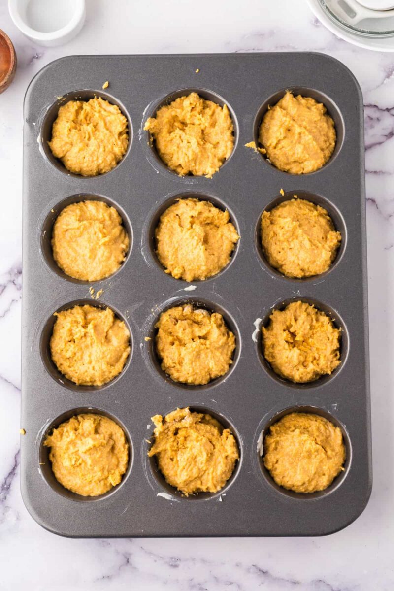 A muffin tin filled with twelve portions of unbaked pumpkin corn muffins batter sits on a white marble countertop.