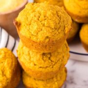 A stack of three golden brown pumpkin corn muffins sits on a marble surface, with more muffins and a bowl of cornmeal in the background. The muffins look moist and slightly crumbly.