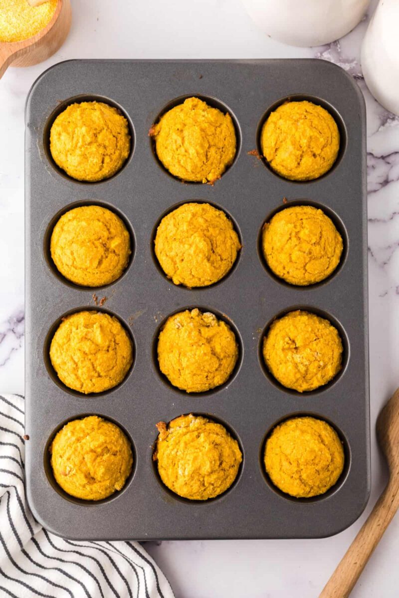 A muffin tin filled with twelve golden-brown pumpkin corn muffins sits on a marble surface, next to a wooden spoon and a striped kitchen towel.