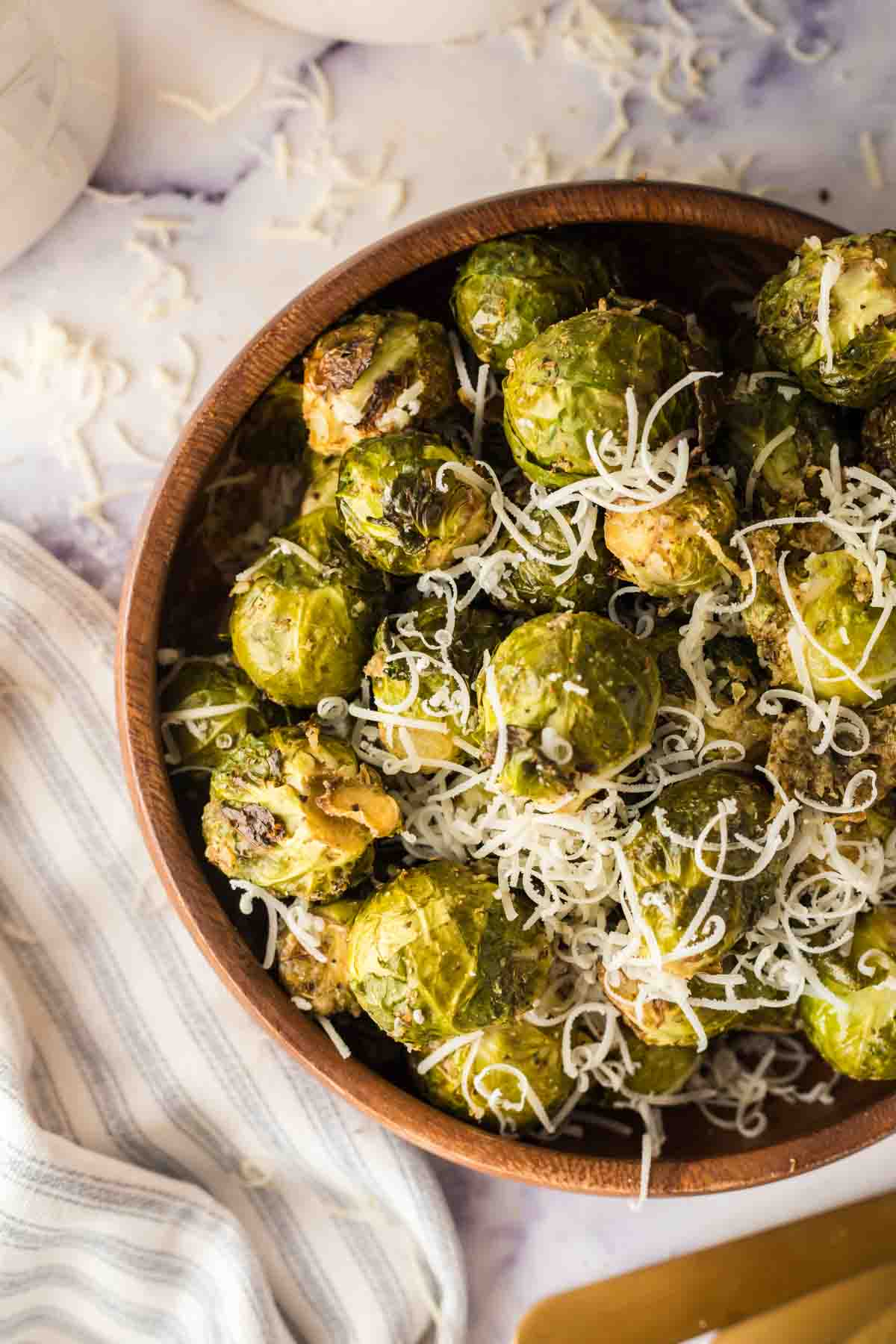 A wooden bowl filled with roasted Brussels sprouts topped with shredded cheese, made using an easy air fryer brussels sprouts recipe, placed on a light-colored surface with a striped cloth nearby.