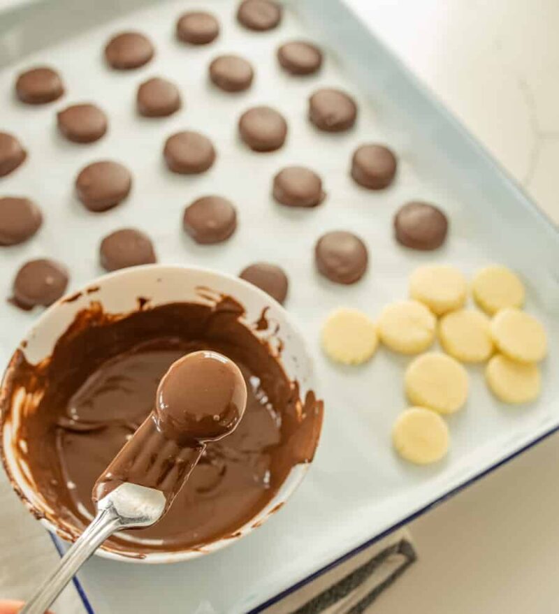 A spoon dips a round, pale treat into melted chocolate in a bowl, just like in a classic peppermint patty recipe. On a parchment-lined tray, chocolate-coated treats and plain, undipped treats are neatly arranged.