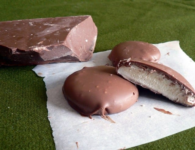 A large block of chocolate and three round chocolate-covered candies with white filling, reminiscent of a peppermint patty recipe, are placed on parchment paper atop a green surface. One candy is cut open, revealing its creamy interior.