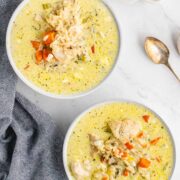 Two bowls of creamy chicken wild rice soup with visible chunks of chicken, carrots, celery, and shredded cheese on top, placed on a white surface next to a blue-gray napkin, two spoons, and a plate of biscuits.