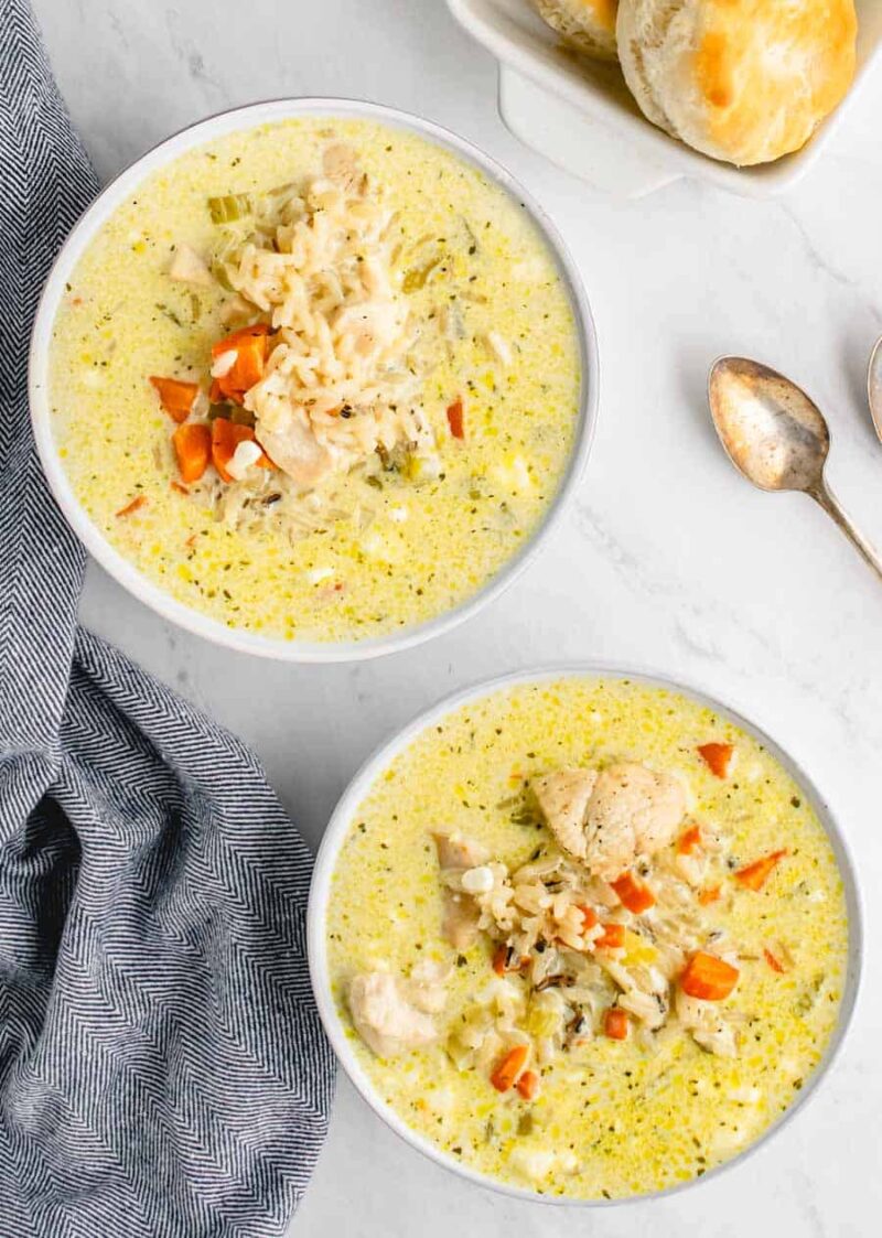 Two bowls of creamy chicken wild rice soup with visible chunks of chicken, carrots, celery, and shredded cheese on top, placed on a white surface next to a blue-gray napkin, two spoons, and a plate of biscuits.