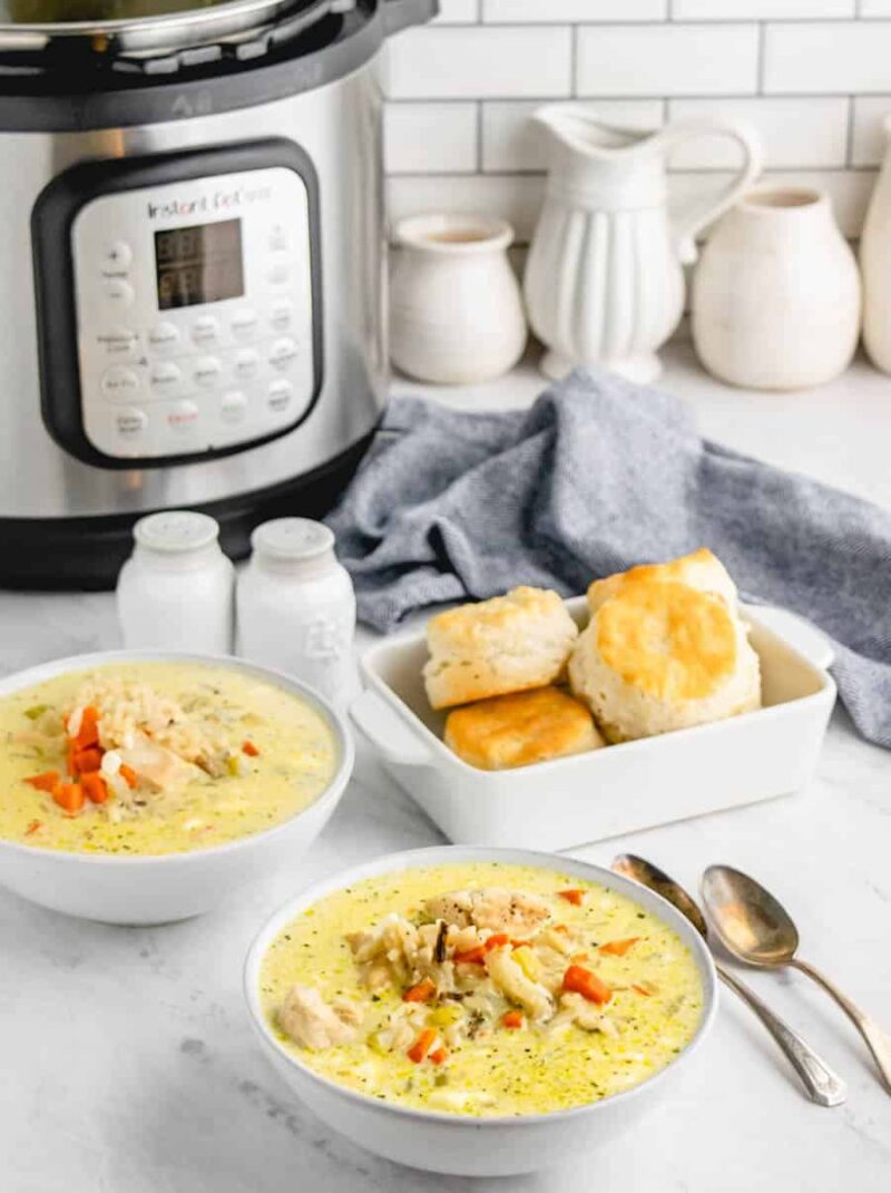 Two bowls of creamy chicken wild rice soup with vegetables sit on a white counter, next to biscuits, two spoons, salt and pepper shakers, a gray napkin, and an Instant Pot in the background.