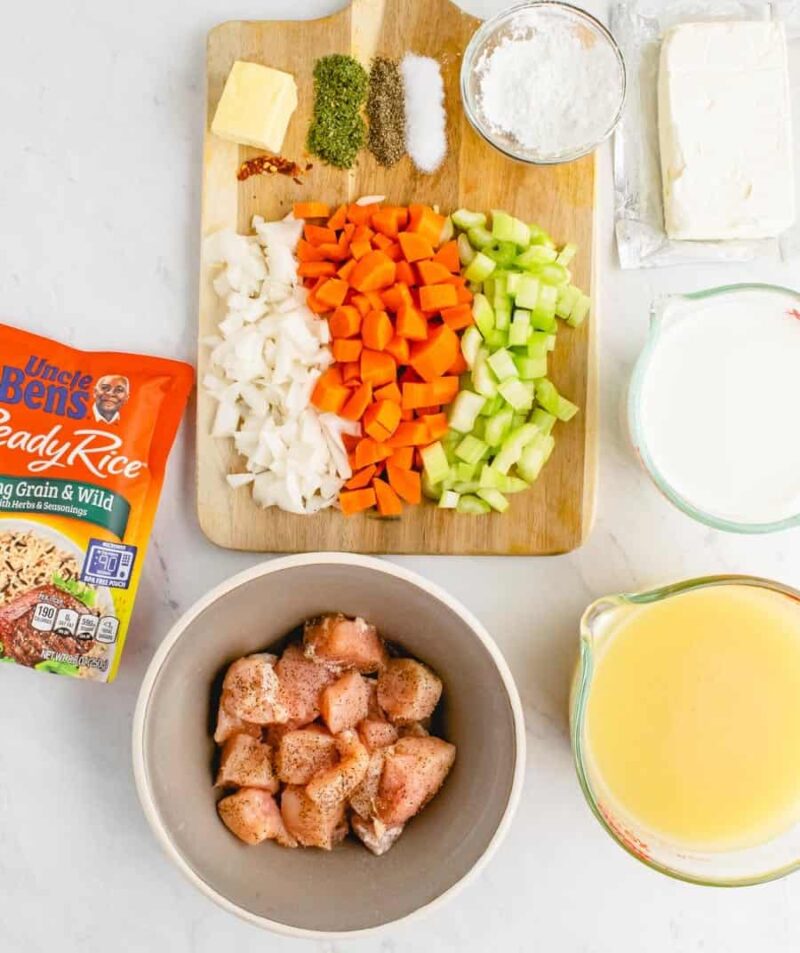 Chopped onion, carrot, and celery on a wooden board with butter, herbs, salt, and flour; bowls hold cream cheese, milk, cooked rice for chicken wild rice soup, and seasoned raw chicken pieces; plus a pitcher of broth.