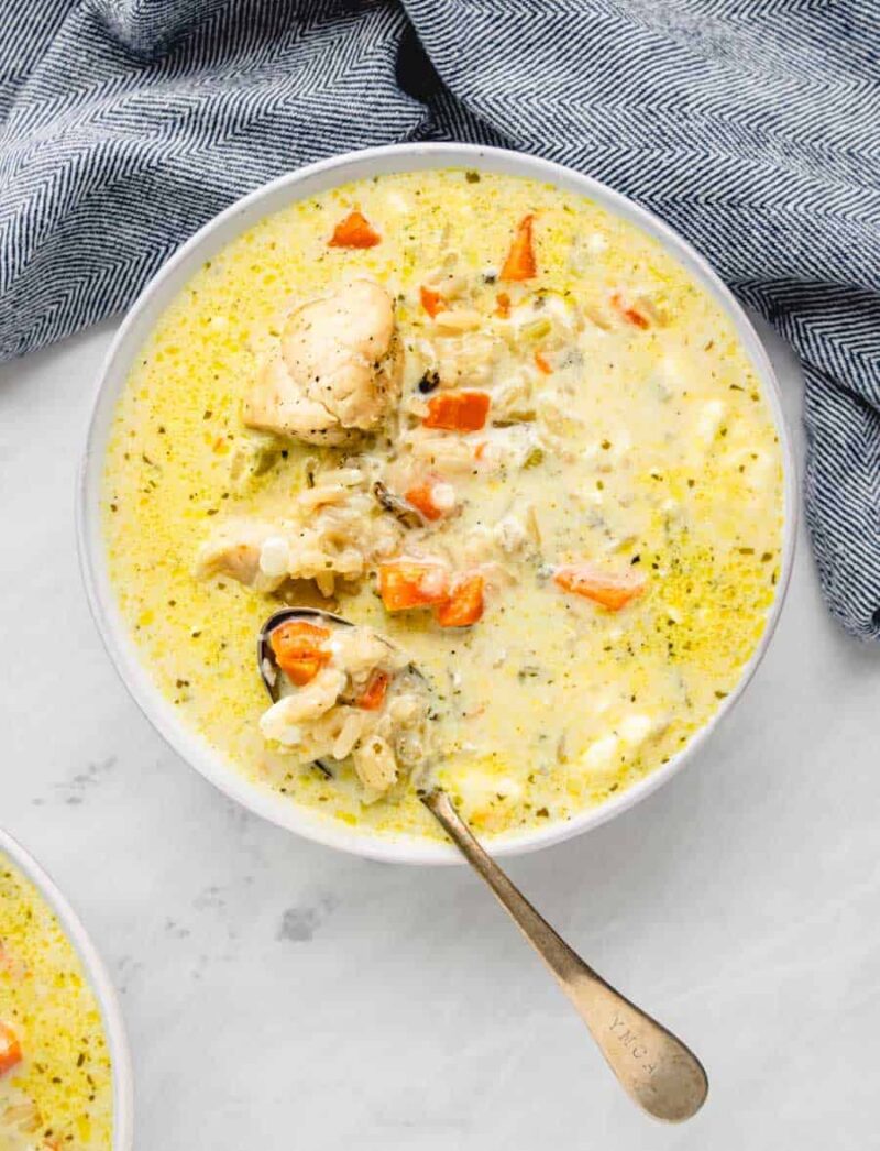 A bowl of creamy chicken wild rice soup with chunks of chicken, carrots, herbs, and wild rice sits alongside a spoon. A blue and white patterned cloth rests nearby on a light surface.