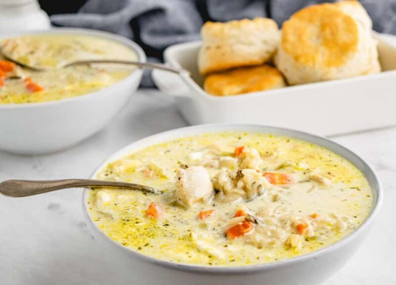 A bowl of creamy chicken wild rice soup with visible carrots and herbs, served with a spoon. In the background, a dish of golden, flaky biscuits. Another bowl of soup is partially visible.