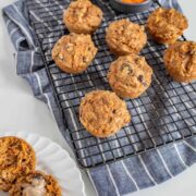 Healthy carrot cake muffins with raisins cool on a rack over a blue striped cloth, alongside a small bowl of grated carrots and a halved muffin with butter melting on a white plate.