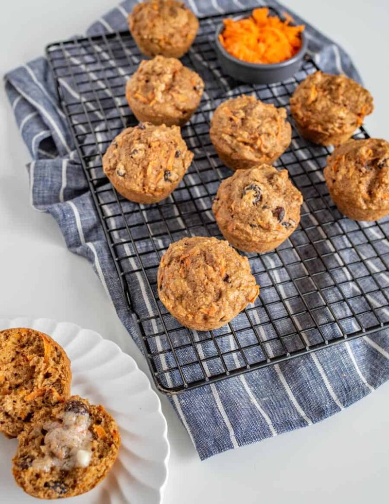 Healthy carrot cake muffins with raisins cool on a rack over a blue striped cloth, alongside a small bowl of grated carrots and a halved muffin with butter melting on a white plate.