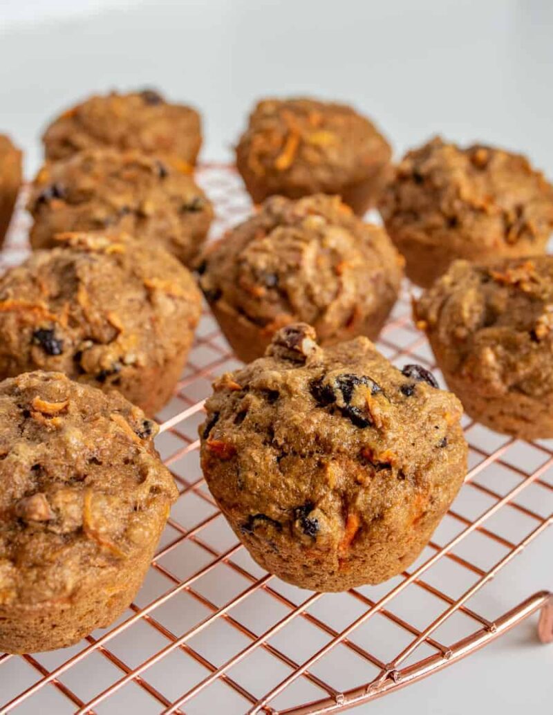 A batch of freshly baked healthy carrot cake muffins with visible pieces of nuts and dried fruit, cooling on a copper wire rack against a light background.