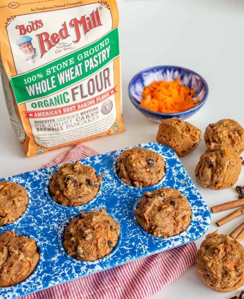 A blue muffin tin with healthy carrot cake muffins, some muffins on the table, a bag of Bob’s Red Mill whole wheat flour, a bowl of grated carrots, cinnamon sticks, and a striped cloth on a white surface.