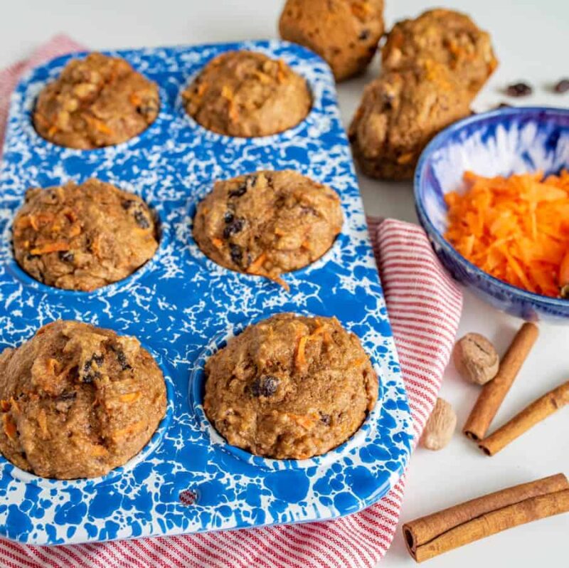 A batch of freshly baked healthy carrot cake muffins with visible pieces of nuts and dried fruit cools on a copper wire rack against a light background.