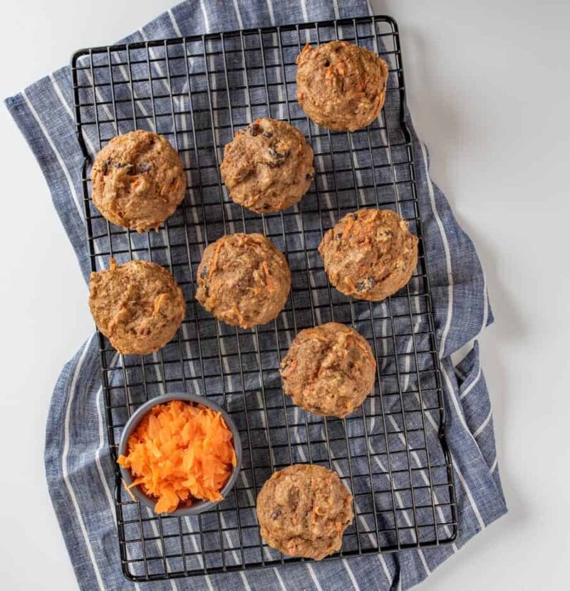 Nine healthy carrot cake muffins are cooling on a black wire rack placed over a blue striped kitchen towel. A small bowl filled with shredded carrots sits among the muffins.