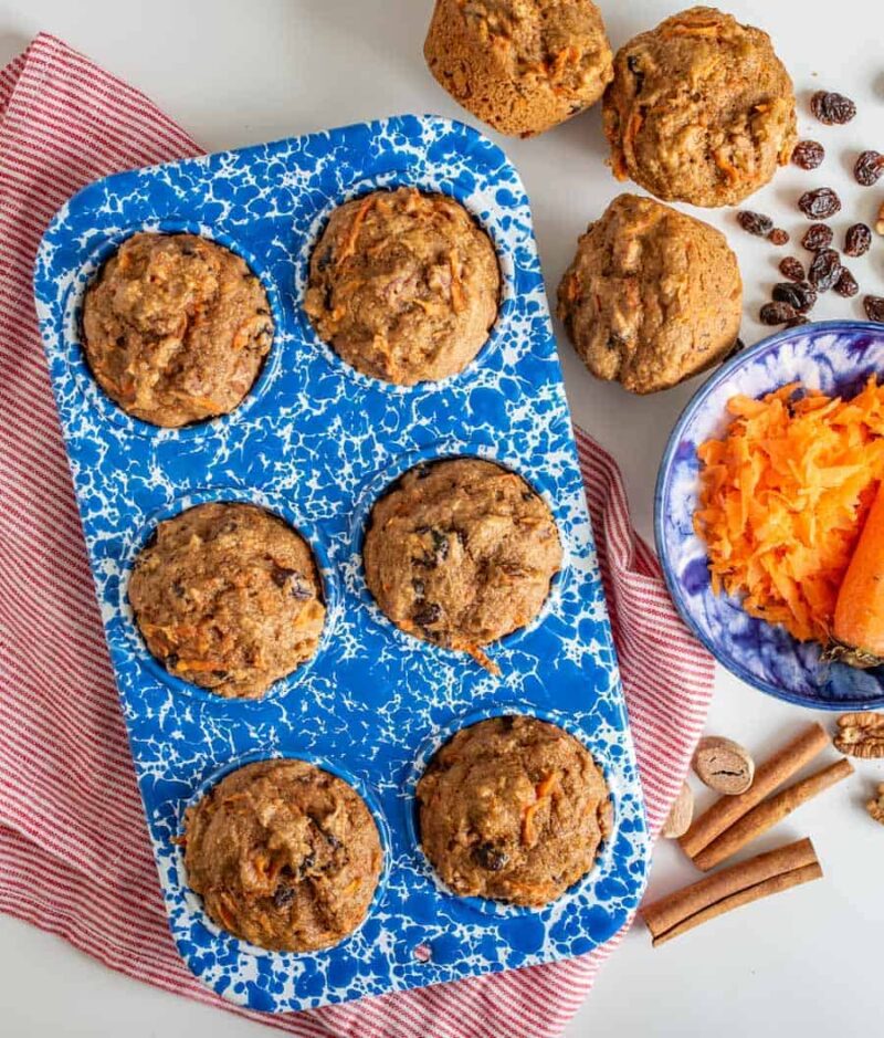 A blue speckled muffin tin holds six baked healthy carrot cake muffins. Nearby are whole muffins, a bowl of shredded carrots with a carrot, cinnamon sticks, walnuts, raisins, and a red-striped cloth on a white surface.