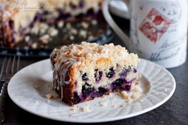 A slice of blueberry crumb cake topped with icing sits on a white plate, with crumbs scattered around and a mug with red and gray designs in the background.