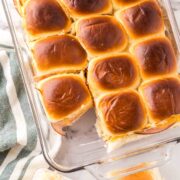 A glass baking dish filled with golden-brown dinner rolls, perfect for a sliders recipe with Hawaiian rolls. One roll is separated and placed on the marble countertop nearby, next to a green and white striped towel.
