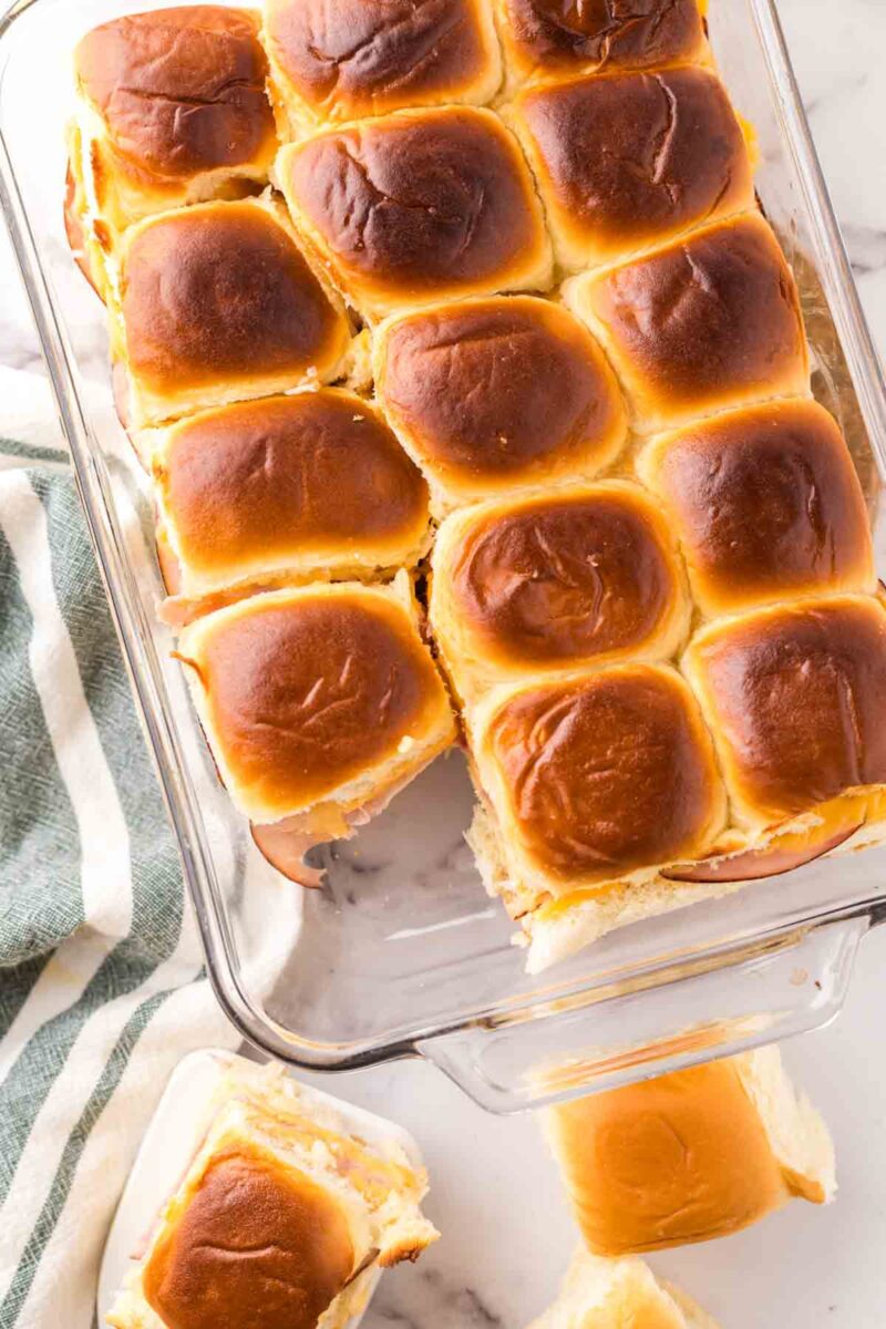 A glass baking dish filled with golden-brown dinner rolls, perfect for a sliders recipe with Hawaiian rolls. One roll is separated and placed on the marble countertop nearby, next to a green and white striped towel.