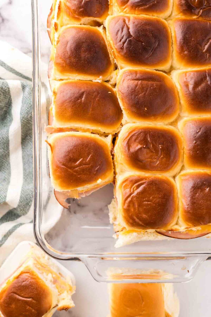 A glass baking dish filled with golden-brown sliders recipe with Hawaiian rolls, some slightly pulled apart to reveal a creamy filling, sits next to a striped kitchen towel. One delicious slider rests on a white plate nearby.
