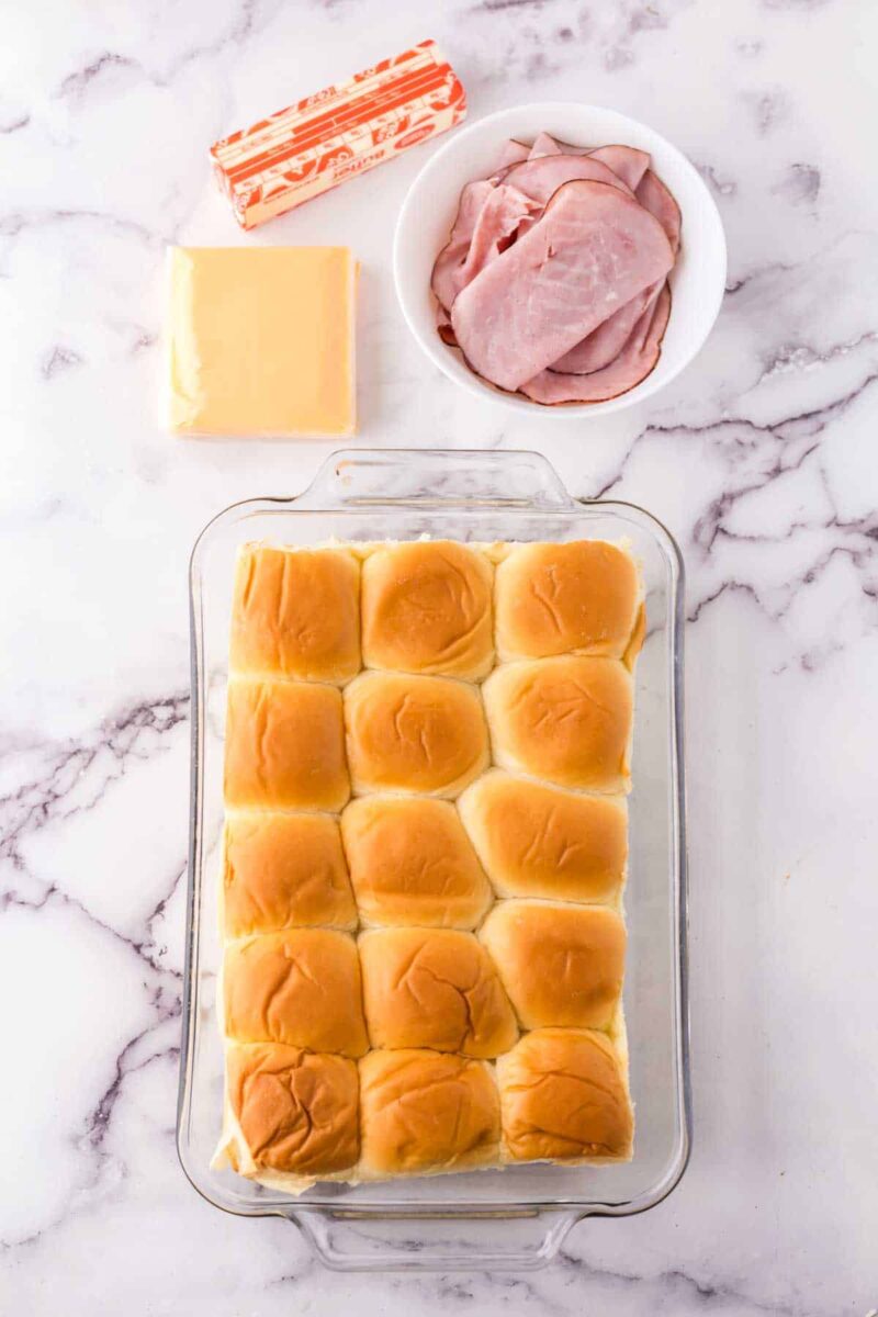 A glass baking dish filled with a sheet of 12 Hawaiian rolls sits on a marble countertop, ready for this tasty sliders recipe with Hawaiian rolls. Nearby are a bowl of sliced ham, cheese slices, and a stick of butter.