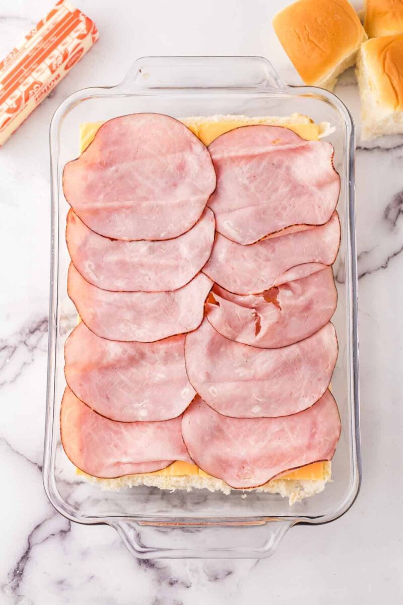 A glass baking dish filled with sliders recipe with Hawaiian rolls, layered with several slices of ham and cheese, with butter and additional rolls nearby on a marble countertop.