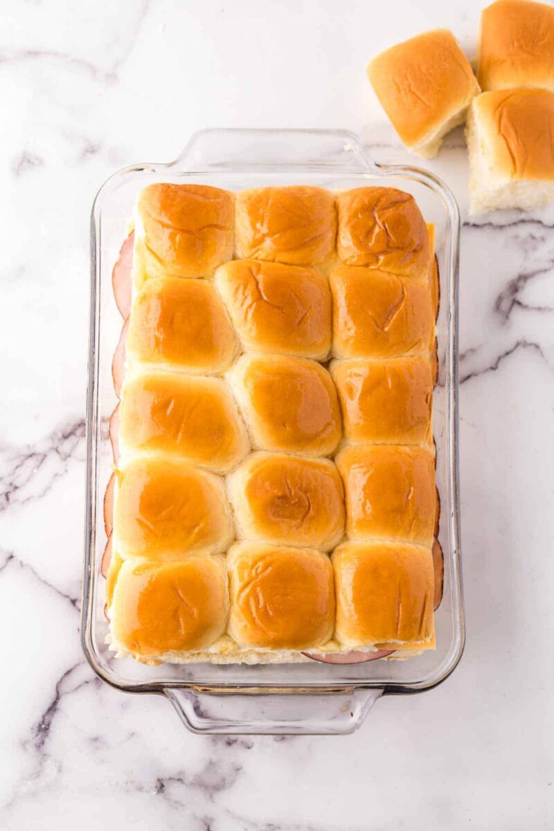 A glass baking dish filled with golden-brown slider rolls, made using a sliders recipe with Hawaiian rolls, sits on a marble surface. Two slider rolls are separated and placed to the upper right of the dish.