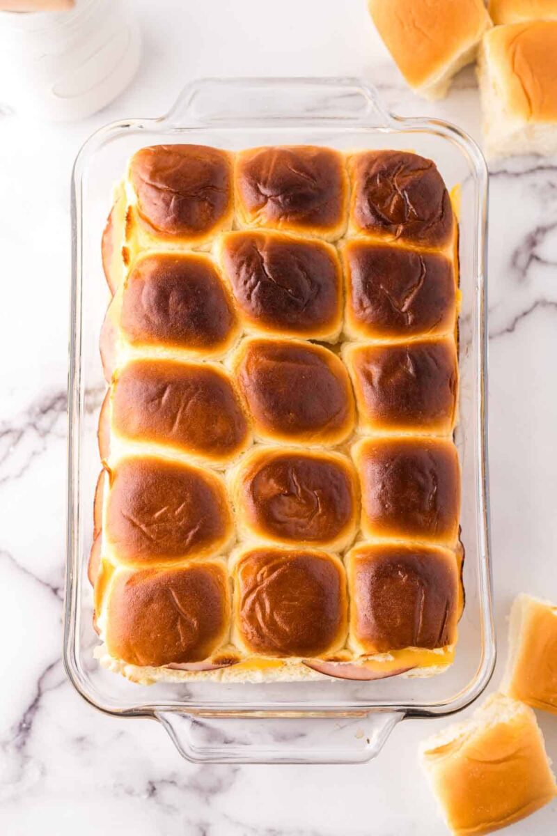A glass baking dish filled with golden-brown, freshly baked Hawaiian rolls sits on a white marble surface—perfect for your next sliders recipe, with a few fluffy rolls removed and placed nearby.