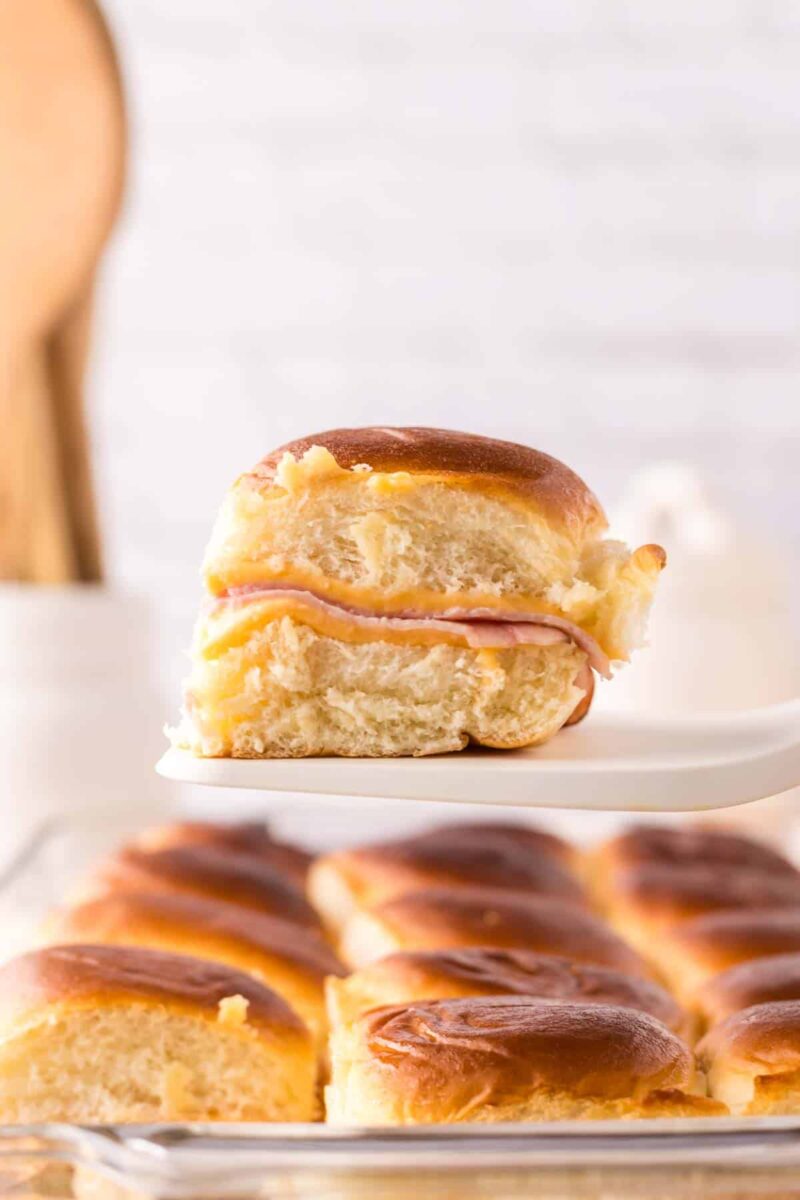 A close-up of a golden-brown slider made with ham and cheese being lifted from a baking dish of similar sandwiches, showcasing a delicious sliders recipe with Hawaiian rolls in a softly blurred kitchen background.