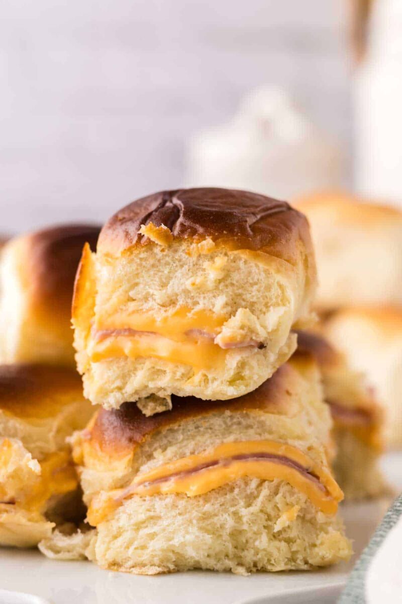 A close-up of two stacked slider sandwiches made with soft, golden-brown Hawaiian rolls filled with melted cheese and deli meat, showcasing a delicious sliders recipe with Hawaiian rolls on a white plate, with more sliders in the background.