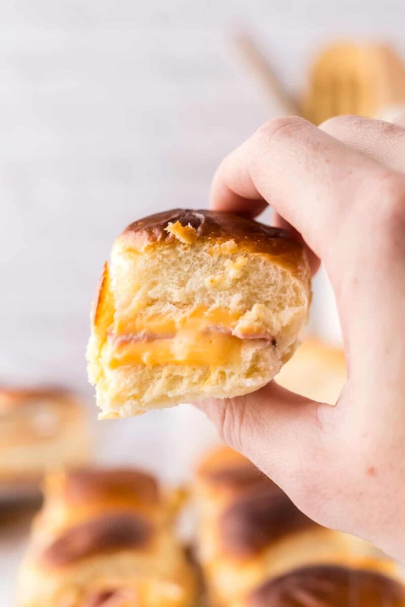 A hand holds a half-eaten soft bread roll with layers of ham and melted cheese inside—a classic sliders recipe with Hawaiian rolls. The background is blurred, showing more freshly baked bread rolls on a tray.