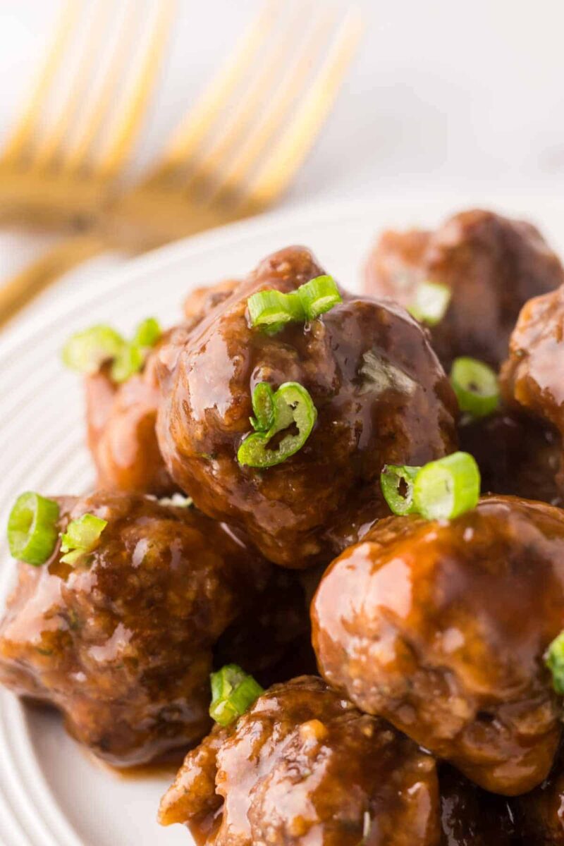 A close-up of sweet and sour meatballs garnished with sliced green onions, served on a white plate with two gold forks blurred in the background.
