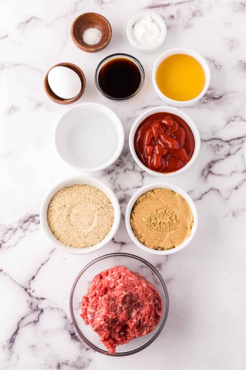 A flat lay of nine ingredients for sweet and sour meatballs in small bowls on a marble surface, featuring ground meat, breadcrumbs, brown sugar, ketchup, melted butter, Worcestershire sauce, an egg, salt, and pepper.