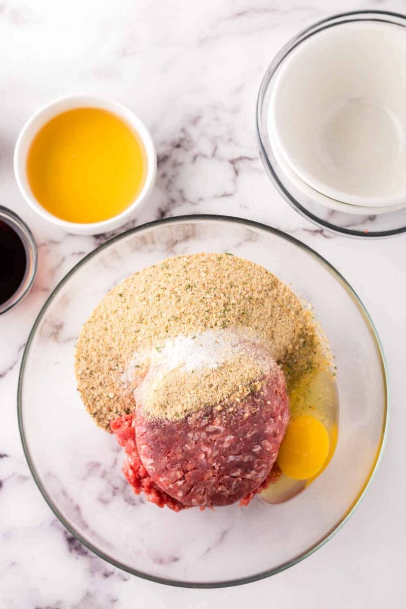 A glass bowl with ingredients for sweet and sour meatballs—ground meat, bread crumbs, an egg, and seasonings—sits on a marble surface alongside bowls filled with yellow liquid and empty white bowls.