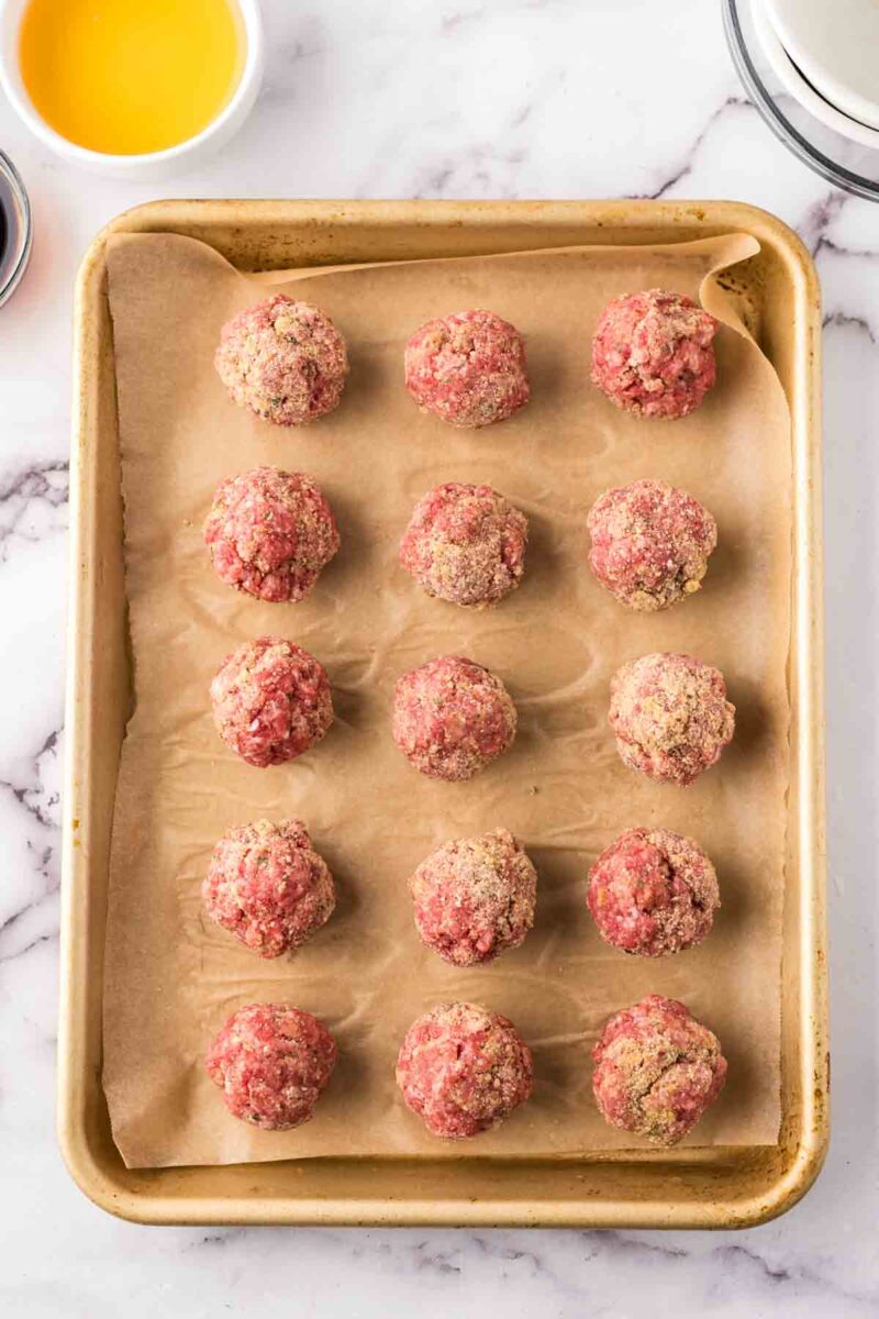 A baking sheet lined with parchment paper holds fifteen raw sweet and sour meatballs evenly spaced in rows. A small bowl with a yellow liquid sits in the upper left corner, all set on a white marble surface.
