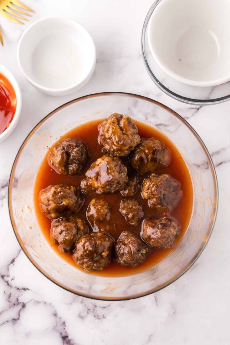 A glass bowl filled with sweet and sour meatballs coated in a reddish-brown sauce sits on a marble countertop, surrounded by empty white bowls and a small dish with red sauce.