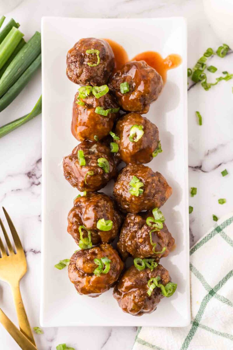 A rectangular white plate holds ten sweet and sour meatballs topped with chopped green onions, arranged neatly on a marble surface. A gold fork, extra green onions, and a green-checked napkin are nearby.