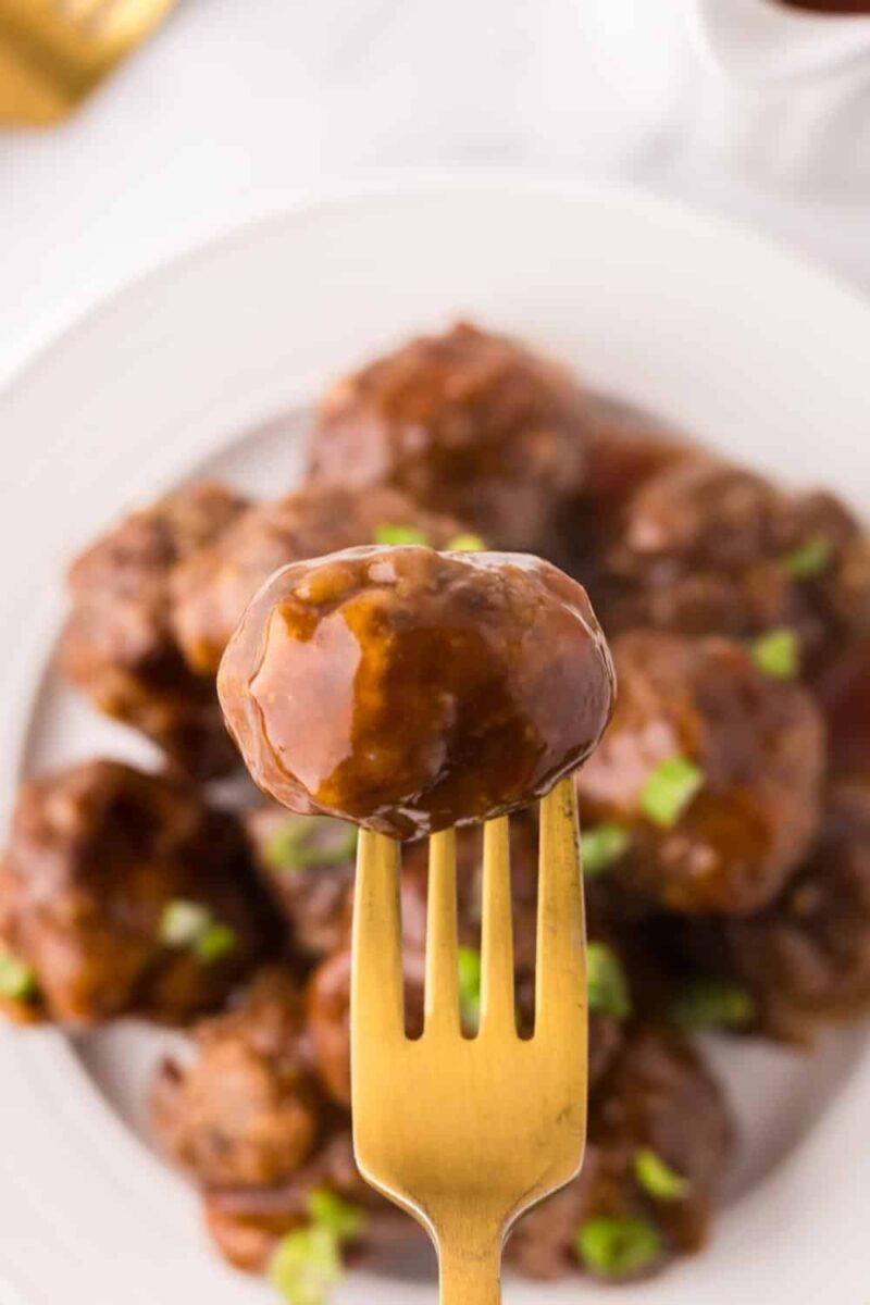 A golden fork holds up a sweet and sour meatball in focus, with a white plate of more glazed sweet and sour meatballs garnished with chopped green onions blurred in the background.