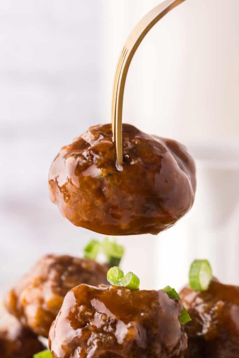 A fork holding a sweet and sour meatball above a plate of more meatballs, all garnished with chopped green onions.