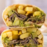 A close-up of a California burrito recipe cut in half, showing layers of guacamole, sliced steak, crispy fries, and melted cheese inside a flour tortilla. The burrito halves are stacked on a white plate.