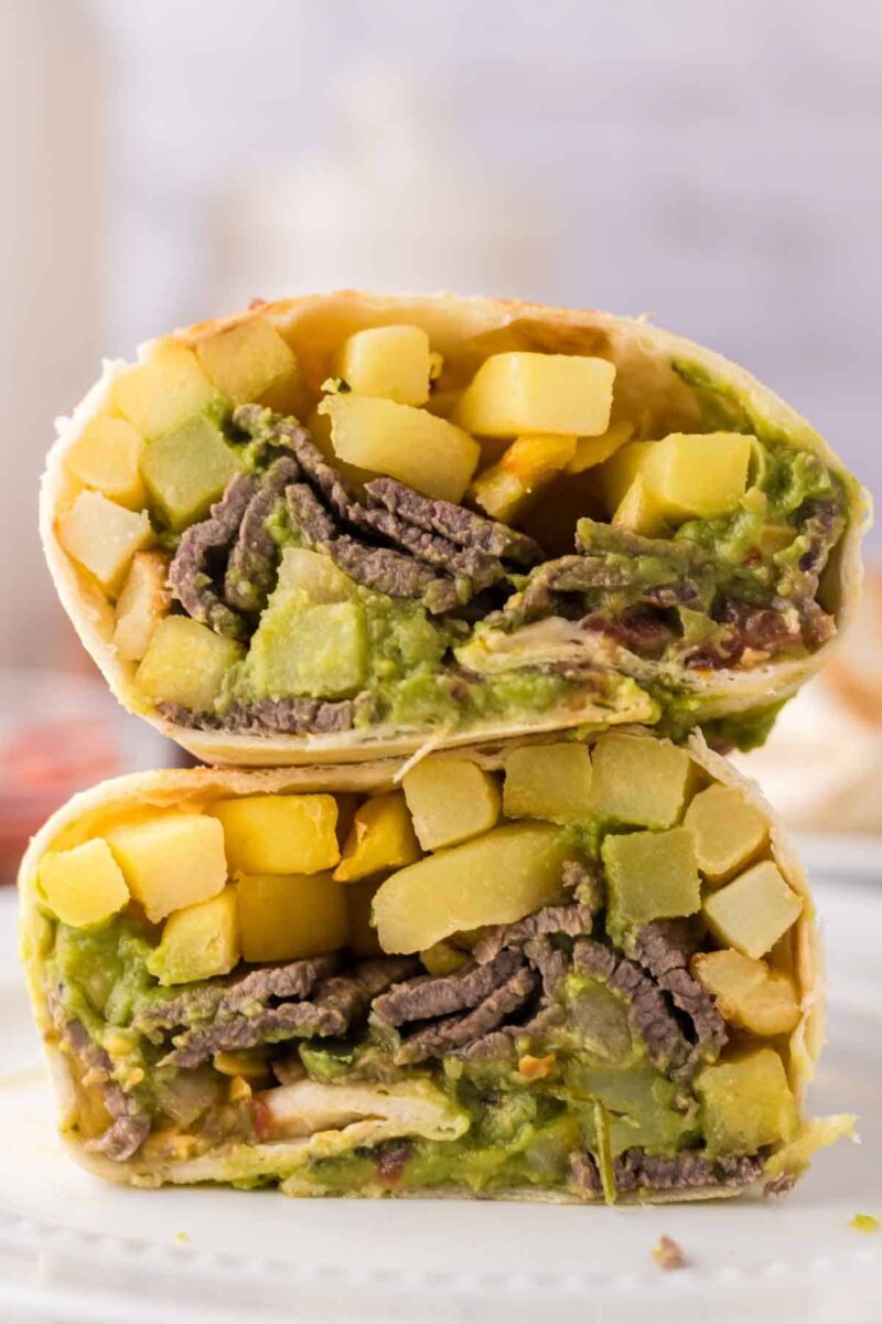 A close-up of a California burrito recipe cut in half, showing layers of guacamole, sliced steak, crispy fries, and melted cheese inside a flour tortilla. The burrito halves are stacked on a white plate.