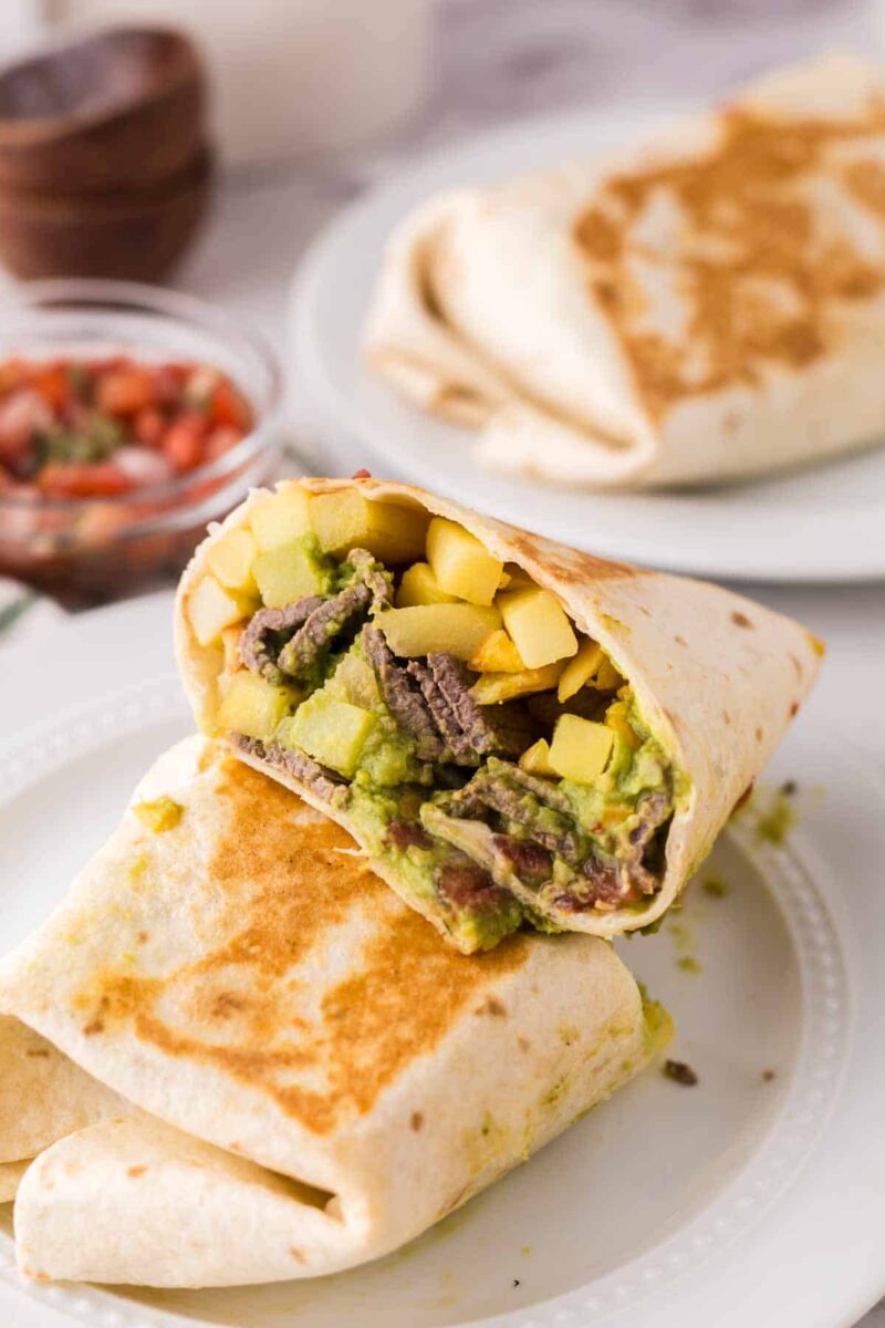 A close-up of a grilled California burrito recipe cut in half, revealing steak, avocado, and vegetables inside, with another whole burrito and a small bowl of salsa in the background.
