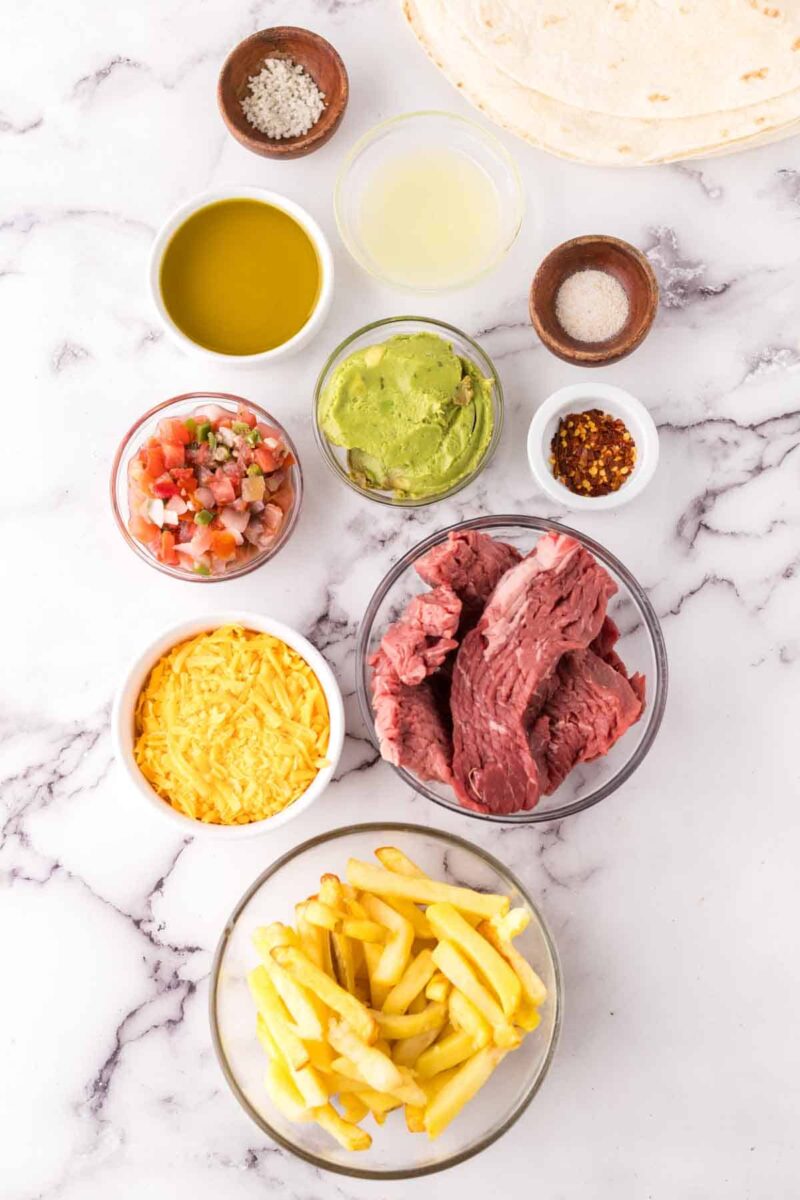 A flat lay of ingredients for a California burrito recipe on a marble surface, featuring raw steak strips, shredded cheddar cheese, French fries, guacamole, pico de gallo, olive oil, lemon juice, tortillas, and various spices in small bowls.