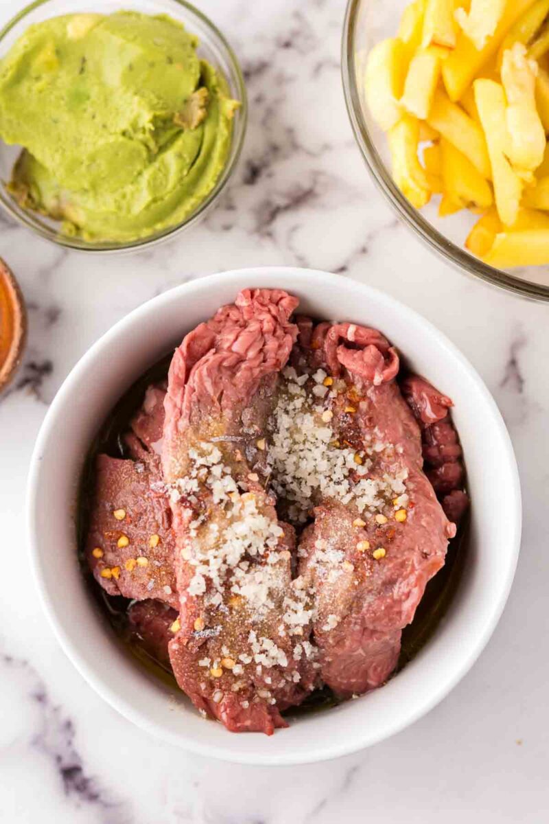 Raw beef strips with seasonings are in a white bowl, ready for a delicious California burrito recipe. Surrounding the bowl are small glass dishes of sliced yellow potatoes or fries and mashed avocado on a white marble surface.