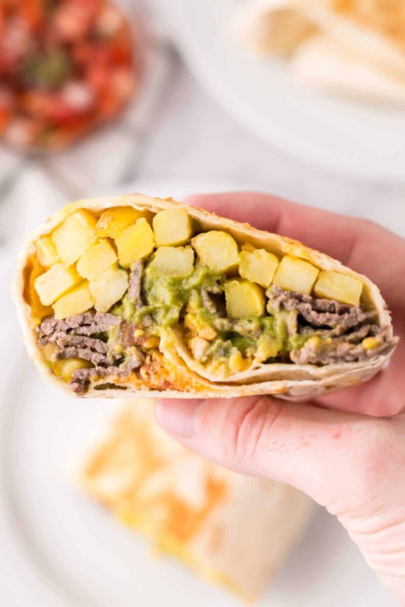 A hand holding a cut burrito, showing layers of golden French fries, seasoned beef, melted cheese, and green sauce inside a soft tortilla—just like a classic California burrito recipe.