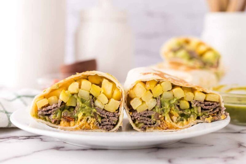 A close-up of a California burrito recipe cut in half, showing layers of steak, crispy fries, melted cheese, and guacamole inside a flour tortilla, on a white plate with another burrito in the background.