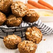 A basket lined with a striped cloth holds several morning glory muffins topped with shredded coconut. Three muffins sit in front on a white surface. In the background are whole carrots and a bowl of pineapple chunks.